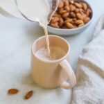 Creamer pouring creamy spiced almond milk with cinnamon and dates into a pink mug with a bowl of almonds behind it.