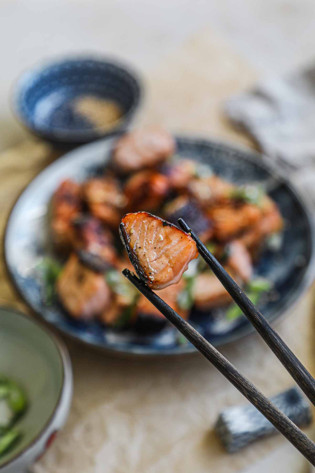 Chopsticks holding honey salmon cubes with sesame seeds and scallions.